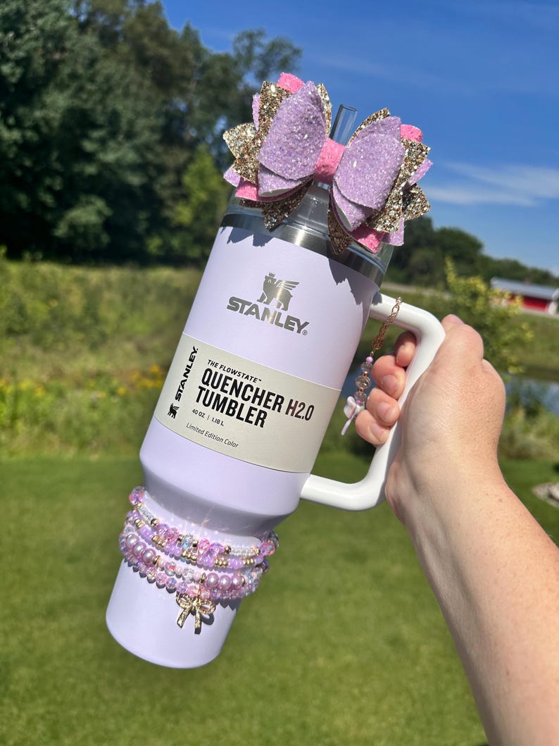 K&ouml;nnte beinhalten: Ein wei&szlig;er Stanley 40 oz (1,18 L) "The Flowstate" Quencher H2.0 Tumbler mit einer Schleife aus lila und goldenen Glitzer oben. Der Becher hat einen Griff und einen Strohhalm. Er ist mit einer Reihe von lila und rosa Perlenarmb&auml;ndern verziert.