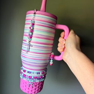 Op de afbeelding: Een roze beker met rietje en handvat, voorzien van horizontale strepen in tinten roze, paars en turquoise. De beker is versierd met diverse kralenbedels, waaronder kleine hart- en strik-hangers die aan het deksel en handvat hangen.