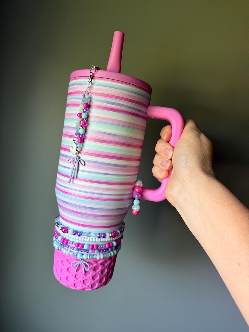 Op de afbeelding: Een roze beker met rietje en handvat, voorzien van horizontale strepen in tinten roze, paars en turquoise. De beker is versierd met diverse kralenbedels, waaronder kleine hart- en strik-hangers die aan het deksel en handvat hangen.