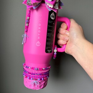 May include: A bright pink HydroJug water bottle with a handle, adorned with a large purple and pink glitter bow on top. The bottle is decorated with beaded bracelets and a beaded charm. The bottle is labeled "HYDROJUG" and "Traveler 32oz".