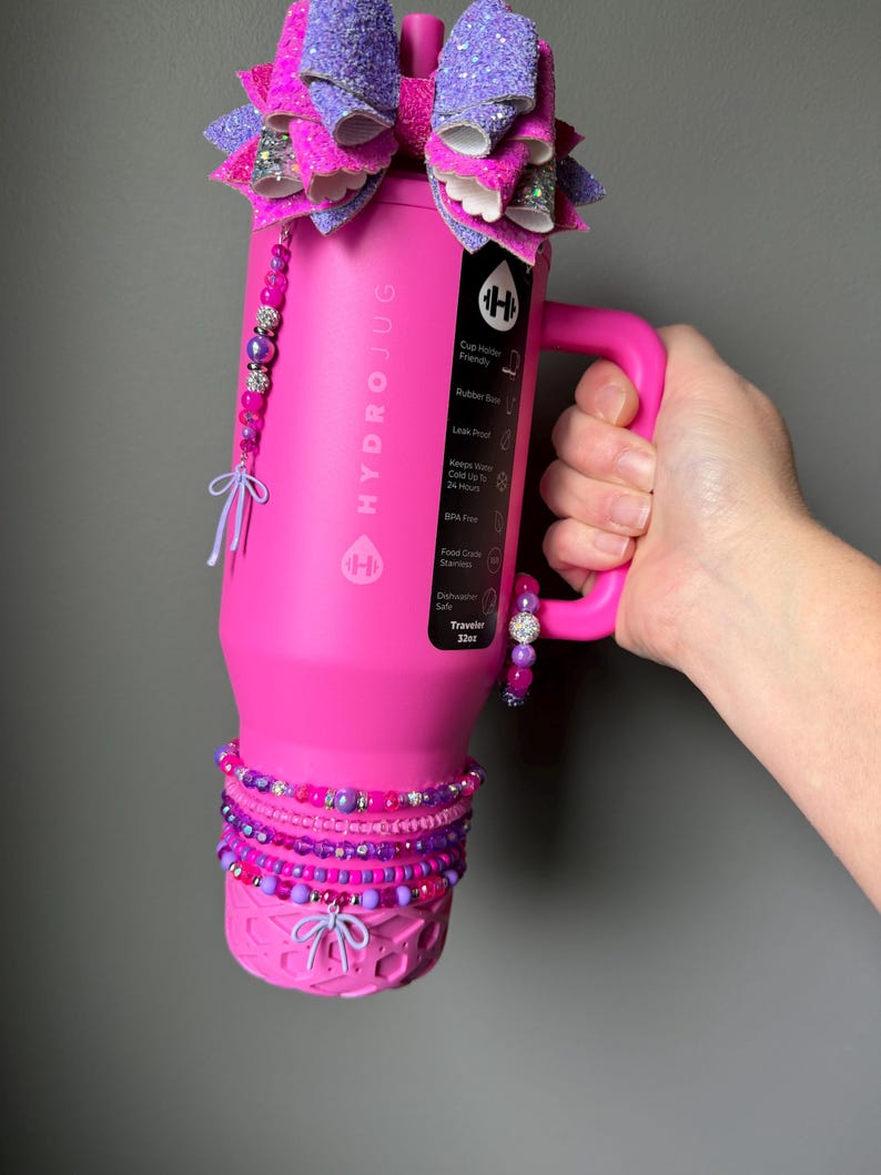 May include: A bright pink HydroJug water bottle with a handle, adorned with a large purple and pink glitter bow on top. The bottle is decorated with beaded bracelets and a beaded charm. The bottle is labeled "HYDROJUG" and "Traveler 32oz".