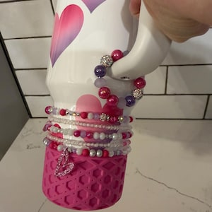 Könnte beinhalten: Ein weiß-rosa isolierter Becher mit Herzdesign. Mehrere Perlenarmbänder in Rosa-, Lila- und Silbertönen sind um den Boden des Bechers gewickelt. Eine rosa Silikonbasis mit Herzanhänger ist ebenfalls sichtbar.