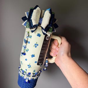 Op de afbeelding: Een cr&egrave;mekleurige 32oz beker met een bosbessenpatroon, een handvat en een blauwe basis met gevlochten textuur. Het deksel is versierd met een grote, meerlaagse glitterstrik in blauwe, groene en witte tinten, geaccentueerd met bungelende kraalbedels.