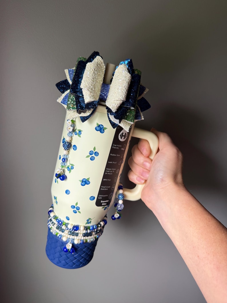 Op de afbeelding: Een cr&egrave;mekleurige 32oz beker met een bosbessenpatroon, een handvat en een blauwe basis met gevlochten textuur. Het deksel is versierd met een grote, meerlaagse glitterstrik in blauwe, groene en witte tinten, geaccentueerd met bungelende kraalbedels.