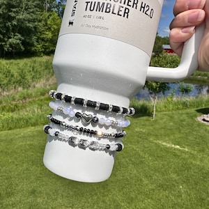 May include: White Stanley Quencher H2.0 Tumbler with black text and a black and white beaded bracelet wrapped around it. The tumbler is 40 oz | 1.18 L and says "All Day Hydration".