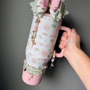 Puede incluir: Un vaso t&eacute;rmico blanco con asa rosa que presenta un patr&oacute;n repetitivo de flamencos rosas, palmeras, coches antiguos y letreros de hotel. El vaso est&aacute; decorado con un gran lazo brillante, abalorios de cuentas y una base rosa texturizada.