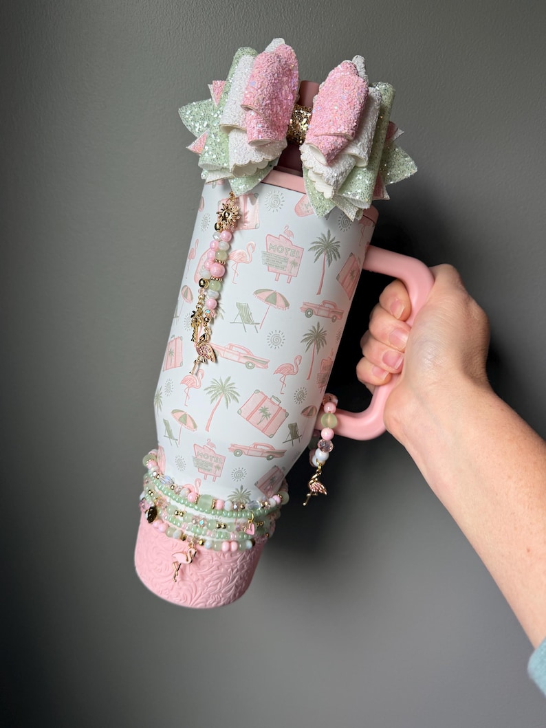 Puede incluir: Un vaso t&eacute;rmico blanco con asa rosa que presenta un patr&oacute;n repetitivo de flamencos rosas, palmeras, coches antiguos y letreros de hotel. El vaso est&aacute; decorado con un gran lazo brillante, abalorios de cuentas y una base rosa texturizada.
