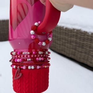 Könnte beinhalten: Eine rote Wasserflasche mit Griff und Armband. Die Flasche hat herzförmige Ausschnitte und eine rote Strickhülle. Die Armbänder sind rot, rosa und silberfarben. Eine Hand hält den Griff.