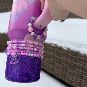 Könnte beinhalten: Eine lila Wasserflasche mit einem rosa Griff und einem Stapel Perlenarmbänder in Rosa-, Lila- und Silbertönen. Die Flasche hat ein Herzdesign und einen dekorativen Sockel mit floralem Muster. Ein silberner Anhänger mit dem Buchstaben "L" hängt an den Armbändern.