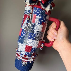 Puede incluir: Un vaso de acero inoxidable con asa roja, que presenta un patr&oacute;n de patchwork en rojo, blanco y azul. La parte superior est&aacute; adornada con un gran lazo brillante, y el cuerpo est&aacute; decorado con dijes de cuentas colgantes y una base azul texturizada.