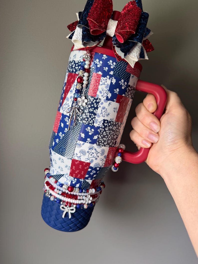 Puede incluir: Un vaso de acero inoxidable con asa roja, que presenta un patr&oacute;n de patchwork en rojo, blanco y azul. La parte superior est&aacute; adornada con un gran lazo brillante, y el cuerpo est&aacute; decorado con dijes de cuentas colgantes y una base azul texturizada.