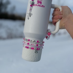May include: White Stanley mug with a pink rim and handle, adorned with beaded bracelets and a charm. The mug features the Stanley logo and is decorated with pink and silver accents. A decorative charm hangs from the top.