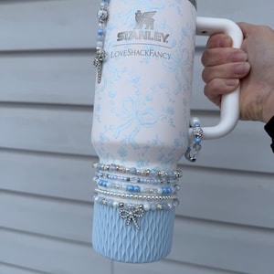 May include: A white and light blue Stanley tumbler with a floral and bow pattern, embellished with beaded bracelets and a decorative bow. The tumbler has a white handle and a light blue base.
