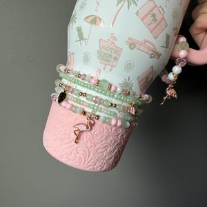 Puede incluir: Un vaso de color rosa claro con un patr&oacute;n tropical de flamencos, palmeras y letreros de motel. El vaso est&aacute; adornado con una pila de pulseras de cuentas en tonos verde claro y rosa, con peque&ntilde;os detalles dorados y un dije de flamenco.