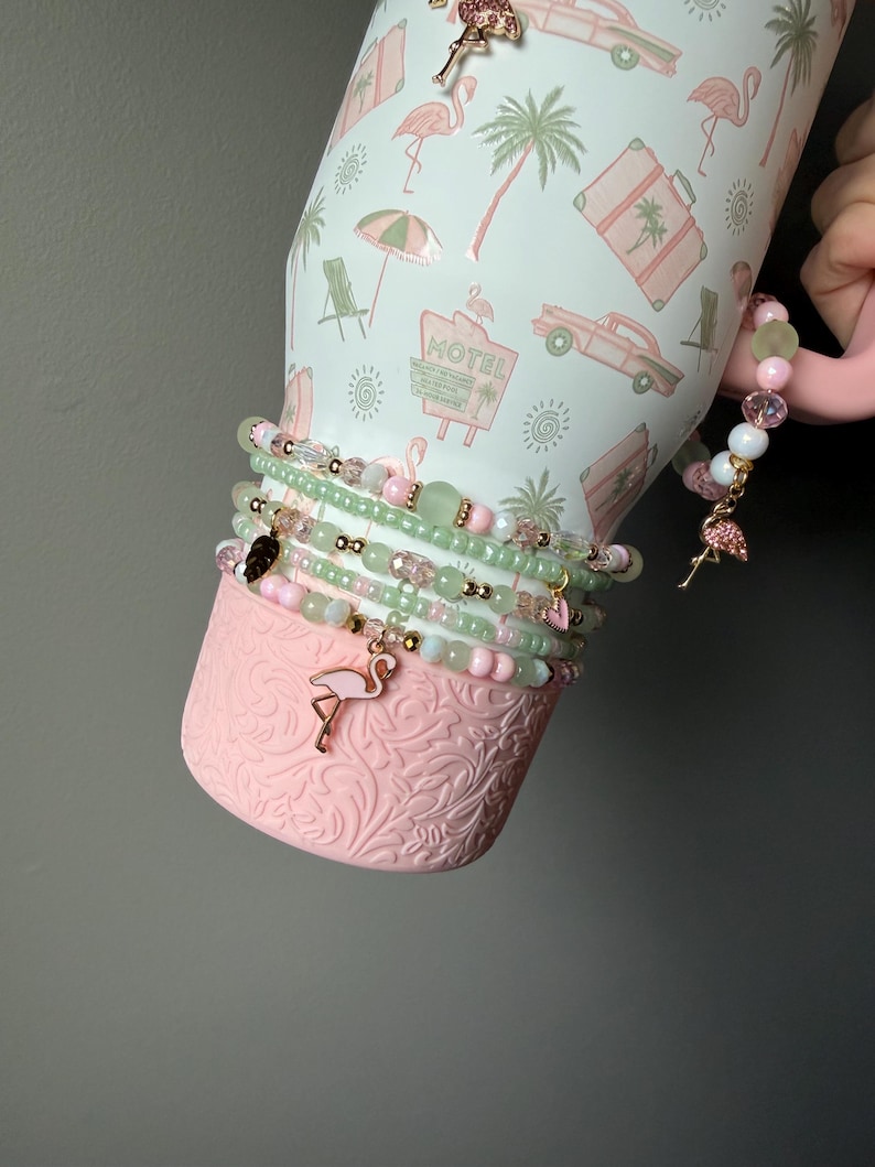Puede incluir: Un vaso de color rosa claro con un patr&oacute;n tropical de flamencos, palmeras y letreros de motel. El vaso est&aacute; adornado con una pila de pulseras de cuentas en tonos verde claro y rosa, con peque&ntilde;os detalles dorados y un dije de flamenco.