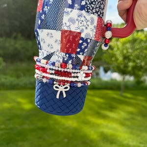 May include: A stainless steel tumbler with a red, white, and blue patchwork pattern, featuring a red handle and a dark blue textured base. The tumbler is adorned with several stacked beaded bracelets in red, white, and blue, including a silver bow charm.