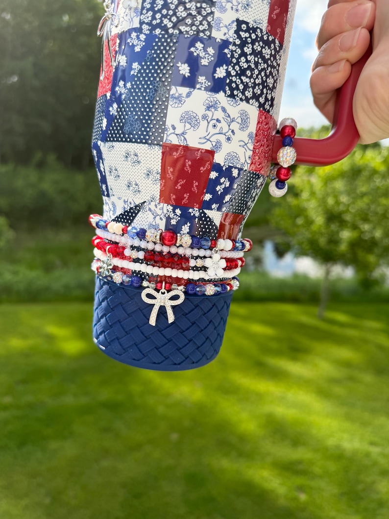 Puede incluir: Un vaso de acero inoxidable con un patr&oacute;n de patchwork en rojo, blanco y azul, con un asa roja y una base texturizada en azul oscuro. El vaso est&aacute; adornado con varias pulseras de cuentas apiladas en rojo, blanco y azul, incluyendo un dije de lazo plateado.