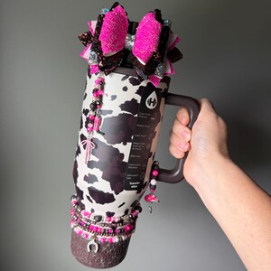 May include: A 40oz stainless steel tumbler with a black and white cow print pattern, featuring a dark brown handle and base. The tumbler is decorated with a large pink and brown glitter bow, beaded charms, and a silver horseshoe charm at the bottom.