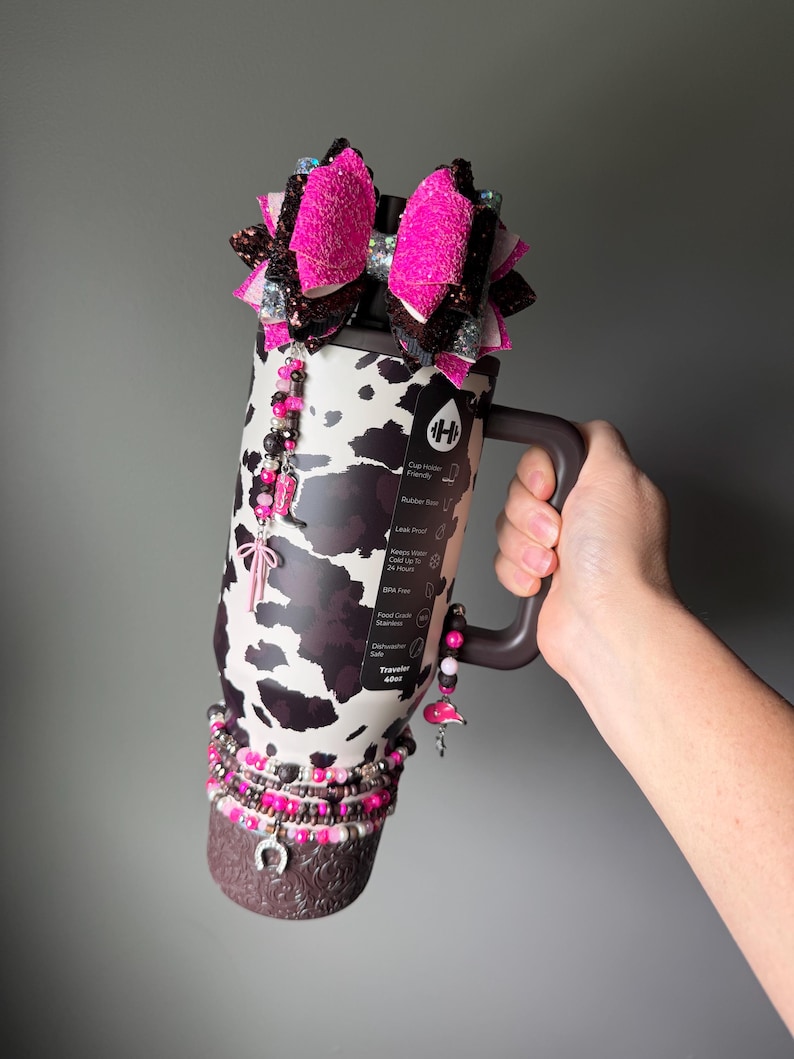 May include: A 40oz stainless steel tumbler with a black and white cow print pattern, featuring a dark brown handle and base. The tumbler is decorated with a large pink and brown glitter bow, beaded charms, and a silver horseshoe charm at the bottom.