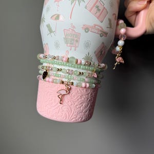 Puede incluir: Un vaso blanco con un patr&oacute;n de motel rosa, palmeras y coches, con una base rosa texturizada. Varias pulseras de cuentas en tonos verde claro y rosa envuelven el vaso, adornadas con peque&ntilde;os dijes de flamencos y corazones.