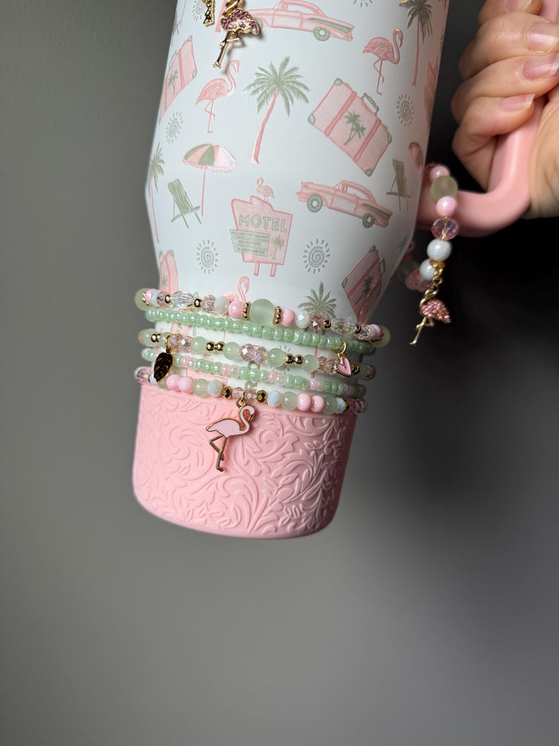 Puede incluir: Un vaso blanco con un patr&oacute;n de motel rosa, palmeras y coches, con una base rosa texturizada. Varias pulseras de cuentas en tonos verde claro y rosa envuelven el vaso, adornadas con peque&ntilde;os dijes de flamencos y corazones.