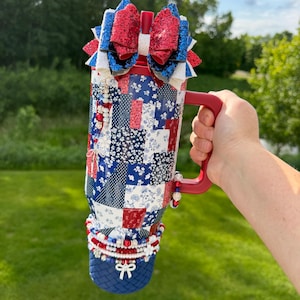 Puede incluir: Un vaso rojo, blanco y azul con un patr&oacute;n de patchwork que presenta dise&ntilde;os florales y geom&eacute;tricos. El vaso tiene un asa roja, una pajita roja y est&aacute; rematado con un gran lazo de purpurina de varias capas. Abalorios de cuentas cuelgan del asa.