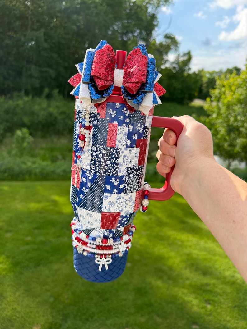 Puede incluir: Un vaso rojo, blanco y azul con un patr&oacute;n de patchwork que presenta dise&ntilde;os florales y geom&eacute;tricos. El vaso tiene un asa roja, una pajita roja y est&aacute; rematado con un gran lazo de purpurina de varias capas. Abalorios de cuentas cuelgan del asa.