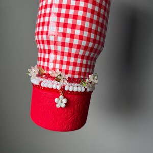K&ouml;nnte beinhalten: Ein rot-wei&szlig; karierter Becher mit floralem Design am Boden. Der Becher ist mit einem dekorativen Band mit wei&szlig;en und rosa Perlen, kleinen Blumen und einem h&auml;ngenden wei&szlig;en Blumenanh&auml;nger verziert.