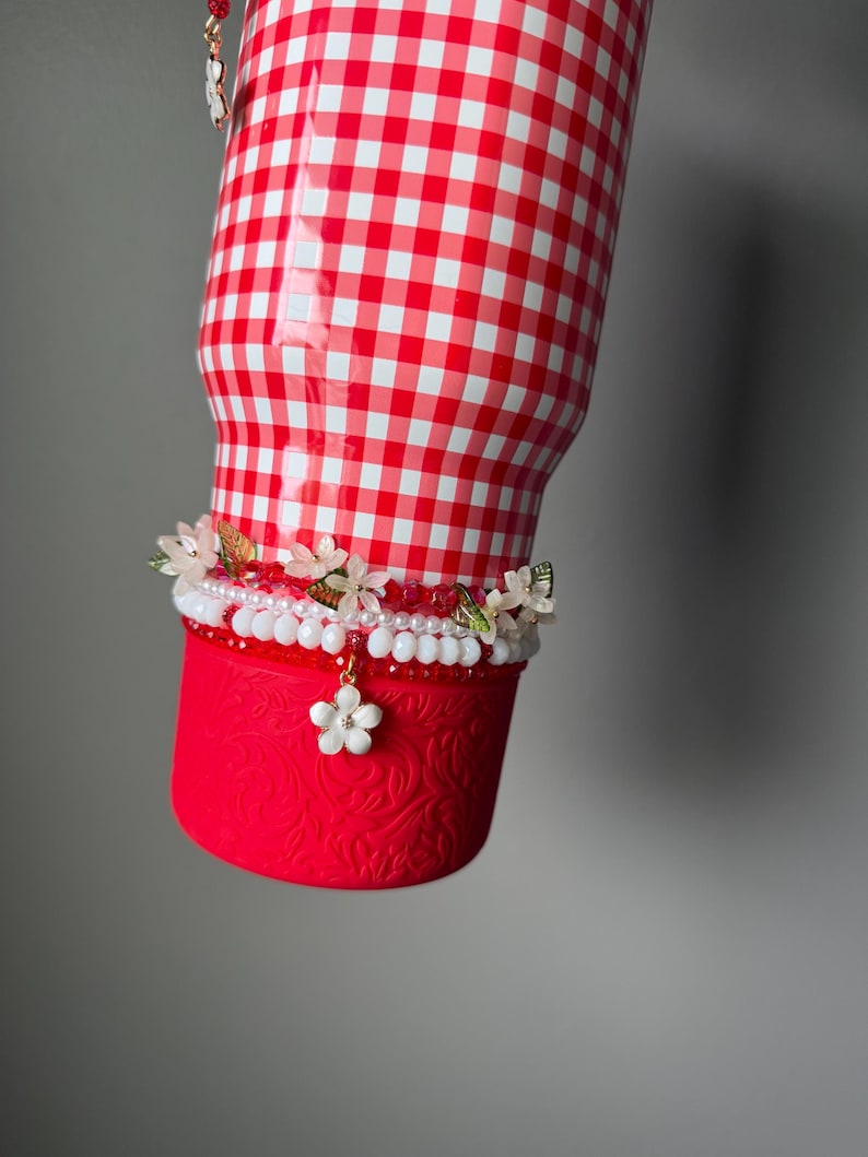 K&ouml;nnte beinhalten: Ein rot-wei&szlig; karierter Becher mit floralem Design am Boden. Der Becher ist mit einem dekorativen Band mit wei&szlig;en und rosa Perlen, kleinen Blumen und einem h&auml;ngenden wei&szlig;en Blumenanh&auml;nger verziert.