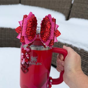 May include: Red Stanley tumbler with a handle, decorated with heart patterns and embellishments. The tumbler is topped with two large, glittery red and pink bows. A beaded charm with hearts hangs from the lid. The tumbler is held by a hand.