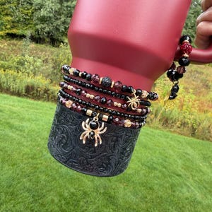 May include: A set of four beaded bracelets featuring black and deep red faceted beads, gold-toned accents, and a prominent gold-toned spider charm with a black center stone. The bracelets are wrapped around a dark red tumbler with a black base.