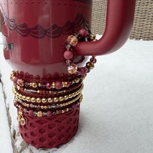 Könnte beinhalten: Ein burgunderfarbener Becher mit einem dekorativen Design und einem Henkel. Mehrere Armbänder mit goldenen und burgunderfarbenen Perlen sind um den Boden und den Henkel des Bechers gewickelt. Der Becher steht auf einer schneebedeckten Oberfläche.