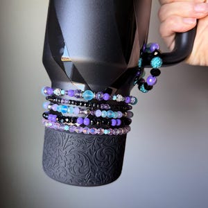 May include: Black insulated tumbler with a geometric design, held by a hand. Several beaded bracelets in shades of purple, black, and teal are stacked around the base of the tumbler. The bracelets feature various bead sizes and textures.