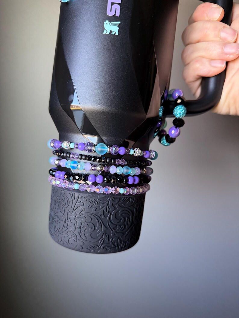 May include: Black insulated tumbler with a geometric design, held by a hand. Several beaded bracelets in shades of purple, black, and teal are stacked around the base of the tumbler. The bracelets feature various bead sizes and textures.