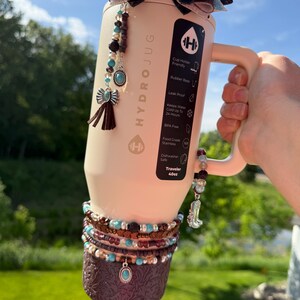 May include: A beige HydroJug water bottle with a handle, decorated with a bow, beaded charms, and a patterned sleeve. The bottle has the text "HYDROJUG" and "Traveler 40oz". The charms include turquoise and brown beads, and a small boot.
