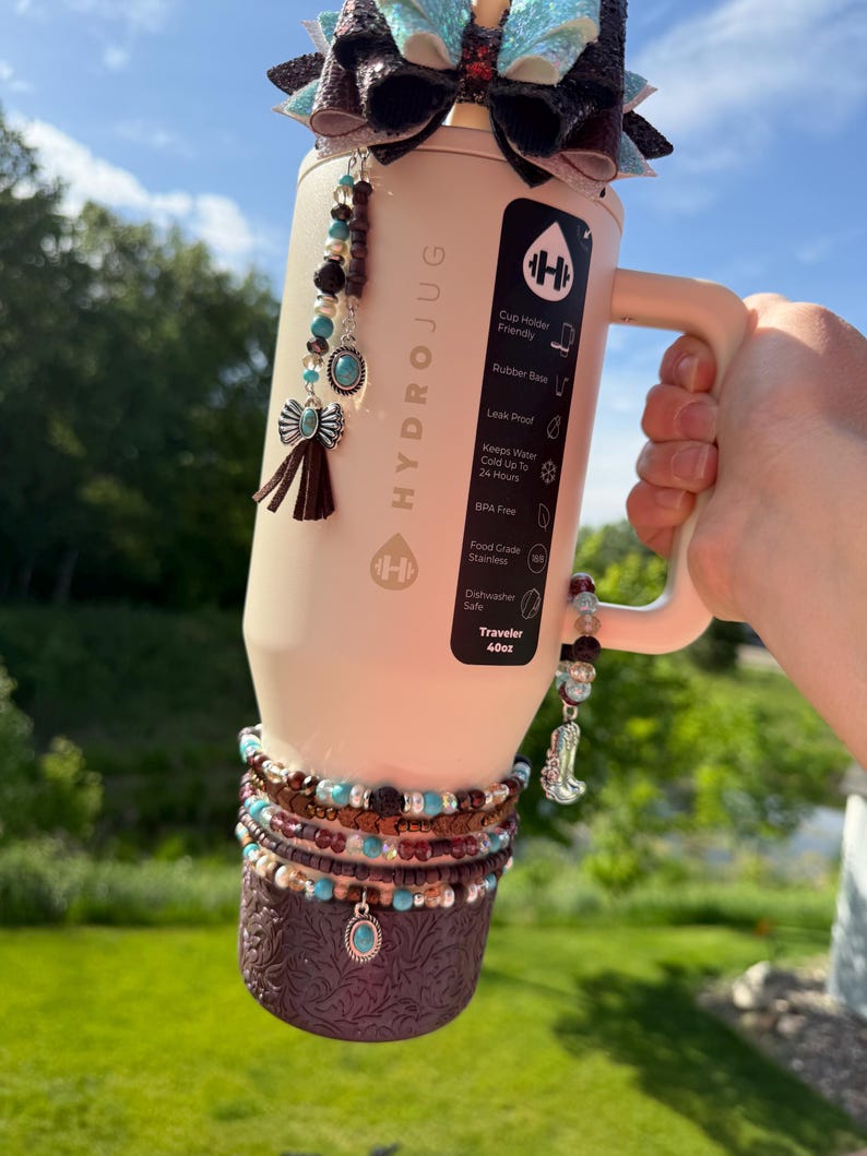 May include: A beige HydroJug water bottle with a handle, decorated with a bow, beaded charms, and a patterned sleeve. The bottle has the text "HYDROJUG" and "Traveler 40oz". The charms include turquoise and brown beads, and a small boot.