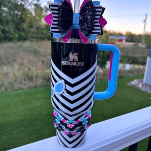 May include: A black and white chevron patterned stainless steel tumbler with a blue and pink handle. The tumbler has a black and white striped bow with glitter on top and a blue and pink beaded bracelet around the middle. The tumbler has the word "STANLEY" printed on the side.