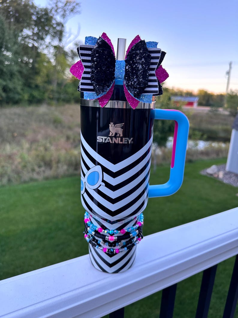 May include: A black and white chevron patterned stainless steel tumbler with a blue and pink handle. The tumbler has a black and white striped bow with glitter on top and a blue and pink beaded bracelet around the middle. The tumbler has the word "STANLEY" printed on the side.