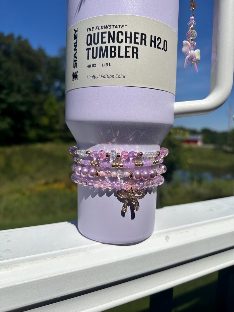 K&ouml;nnte beinhalten: Ein lilafarbener Edelstahlbecher mit einem wei&szlig;en Etikett, auf dem "The Flowstate Quencher H2.0 Tumbler 40 oz | 1,18 L Limited Edition Color" steht. Der Becher ist mit einem Stapel rosafarbener und goldener Perlenarmb&auml;nder verziert.
