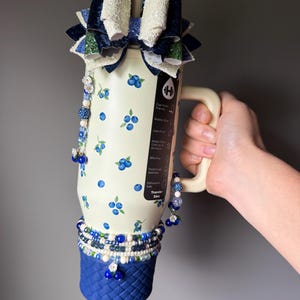 Op de afbeelding: Een 946 ml cr&egrave;mekleurige roestvrijstalen beker met een bosbessenpatroon, een blauwe basis met gevlochten textuur, een grote meerlaagse glitterstrik als bovenkant en bungelende kralenbedels in blauwe, witte en zilveren tinten.