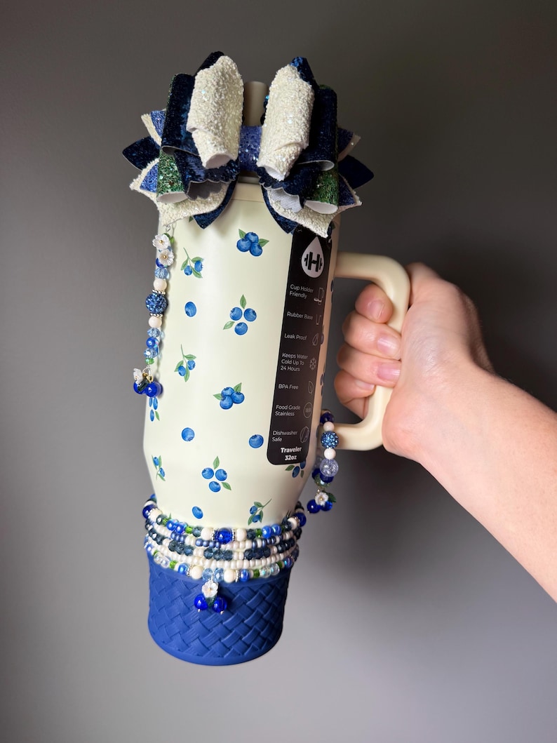 Op de afbeelding: Een 946 ml cr&egrave;mekleurige roestvrijstalen beker met een bosbessenpatroon, een blauwe basis met gevlochten textuur, een grote meerlaagse glitterstrik als bovenkant en bungelende kralenbedels in blauwe, witte en zilveren tinten.