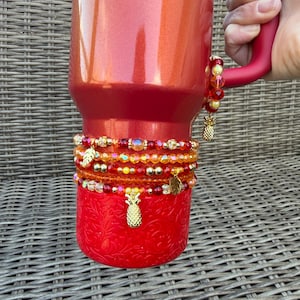 May include: A red tumbler with gold pineapple charms and beaded bracelets in shades of red, orange, and gold. The tumbler has a textured base and a red handle. The bracelets are adorned with gold beads and charms.
