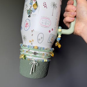 May include: A white travel mug with a green handle and base, adorned with watercolor illustrations of palm trees and floral designs. Several beaded bracelets in shades of blue, green, and yellow are wrapped around the mug, accented with charms.