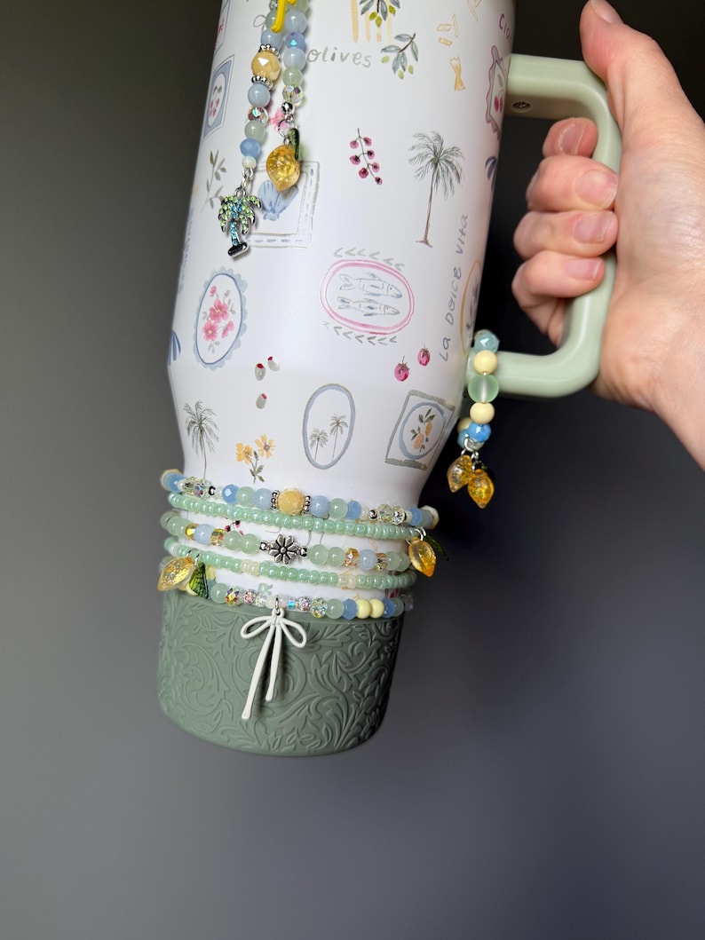 May include: A white travel mug with a green handle and base, adorned with watercolor illustrations of palm trees and floral designs. Several beaded bracelets in shades of blue, green, and yellow are wrapped around the mug, accented with charms.
