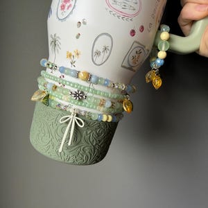 May include: A white ceramic mug with floral and palm tree illustrations, adorned with several beaded bracelets in shades of green, blue, and yellow. The bracelets feature charms and decorative beads. The mug has a green handle and base.