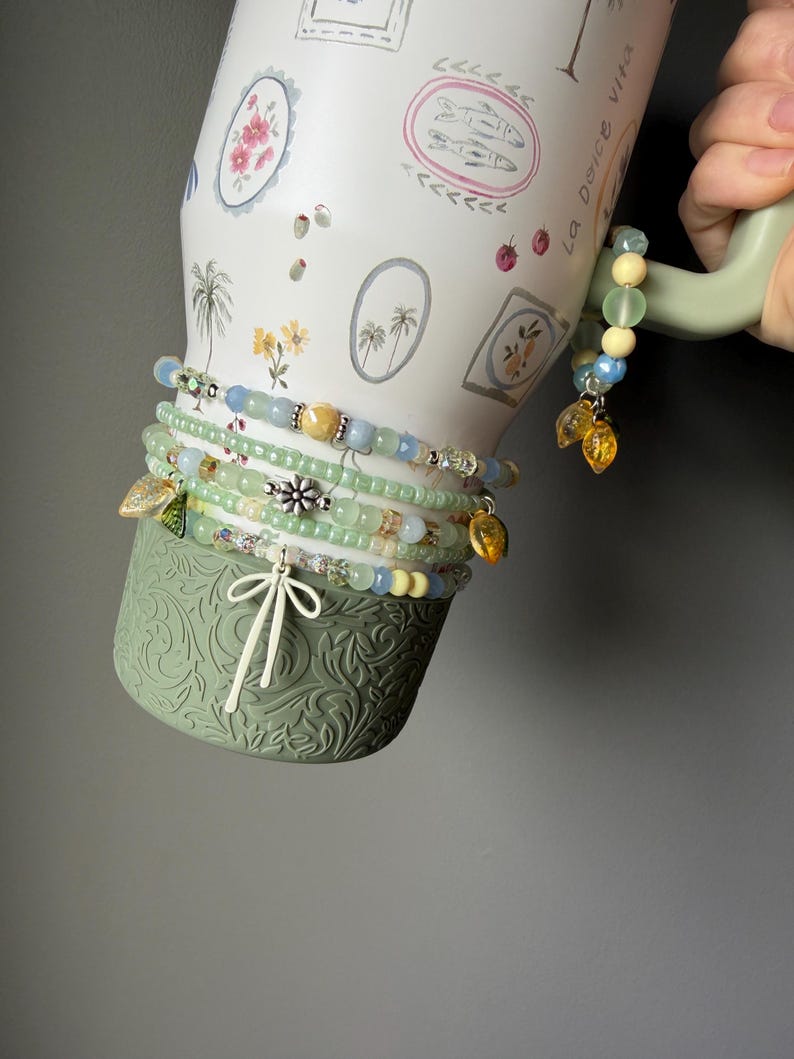 May include: A white ceramic mug with floral and palm tree illustrations, adorned with several beaded bracelets in shades of green, blue, and yellow. The bracelets feature charms and decorative beads. The mug has a green handle and base.