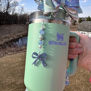 May include: A light green stainless steel travel mug with a white and purple bear logo that says "STANLEY". The mug has a silver lid with a light blue and silver glitter bow attached to the top. A silver chain with light blue and silver beads hangs from the lid with a light blue glitter bow at the bottom.