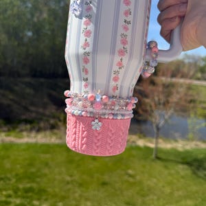 May include: A floral-patterned tumbler with pink and white stripes, adorned with beaded bracelets and charms. The bottom features a pink knitted cozy. The tumbler is held by a person.