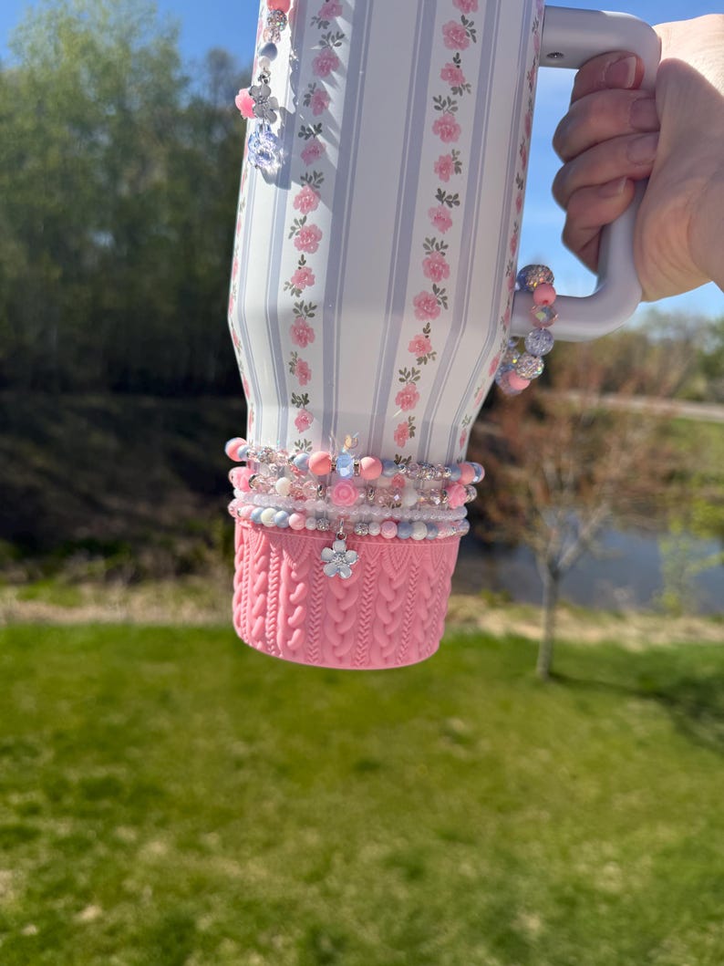 May include: A floral-patterned tumbler with pink and white stripes, adorned with beaded bracelets and charms. The bottom features a pink knitted cozy. The tumbler is held by a person.