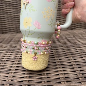 May include: A light green Stanley tumbler with a floral design in pink and yellow. The tumbler is adorned with beaded bracelets in shades of pink, yellow, and green. A gold charm with a pink flower hangs from the bottom.